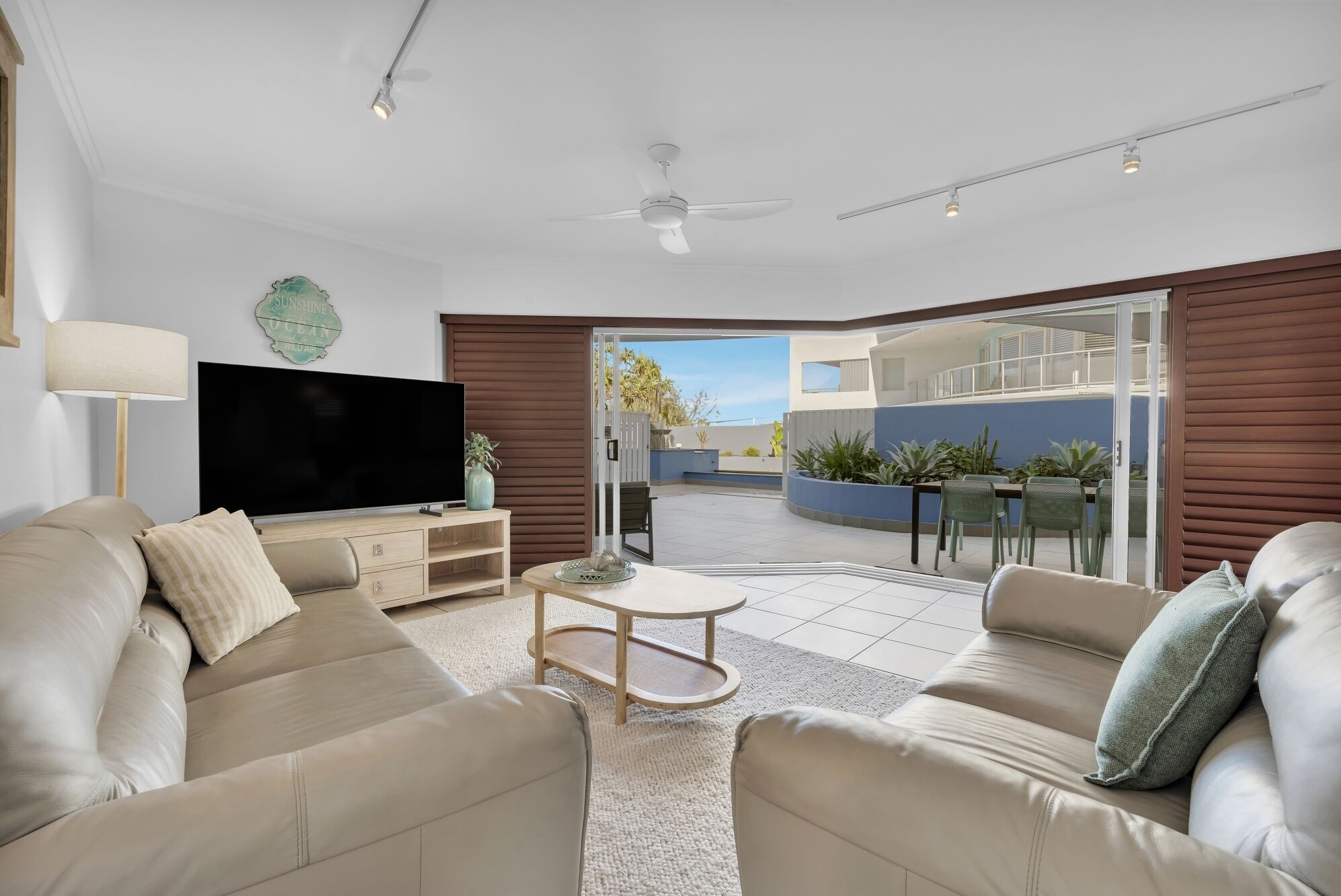 Una sala moderna con una televisión de pantalla plana, sofás color beige, una mesa de centro y un ventanal corredero que da a un patio exterior con una mesa y sillas de comedor.