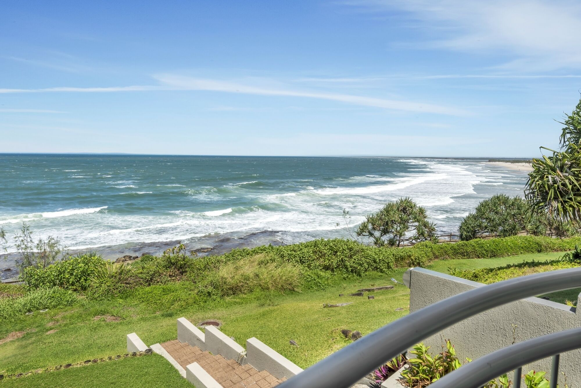 A coastal view with a grassy area, a concrete pathway, and a metal railing.