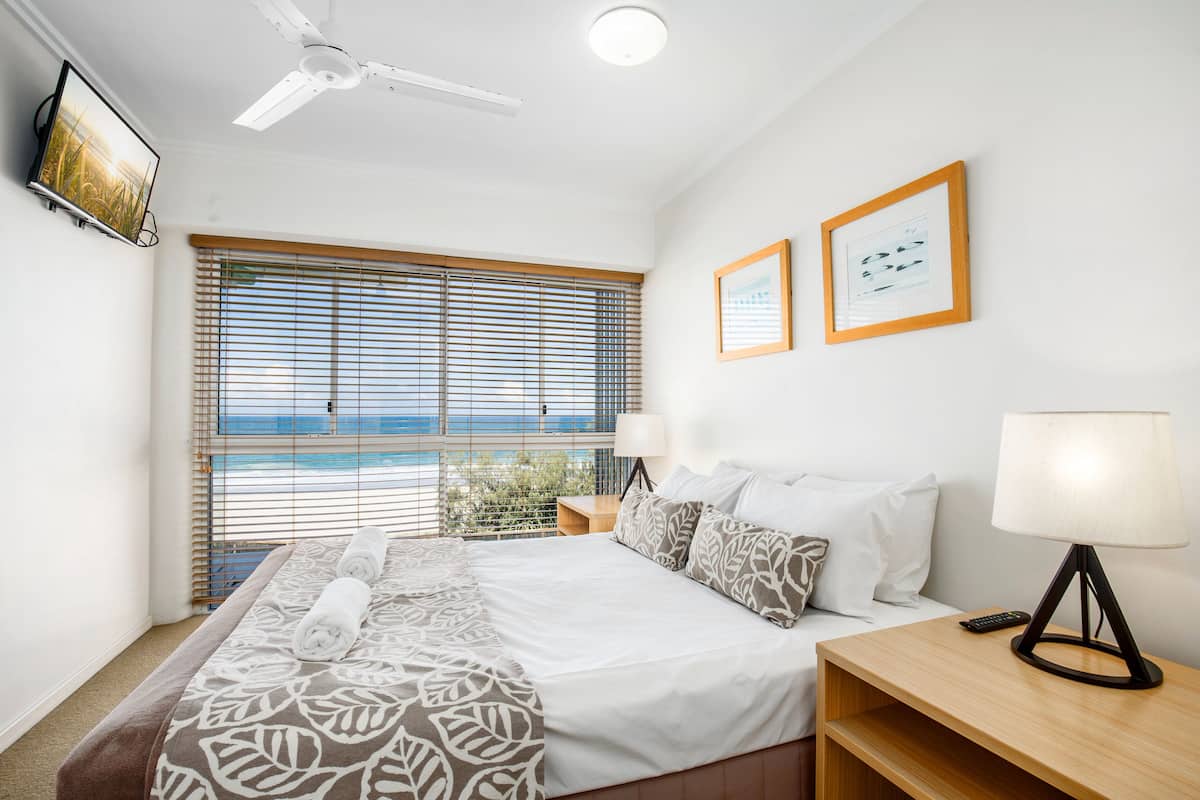 A hotel room with a bed, a bedside table with a lamp, a ceiling fan, and a large window with blinds.
