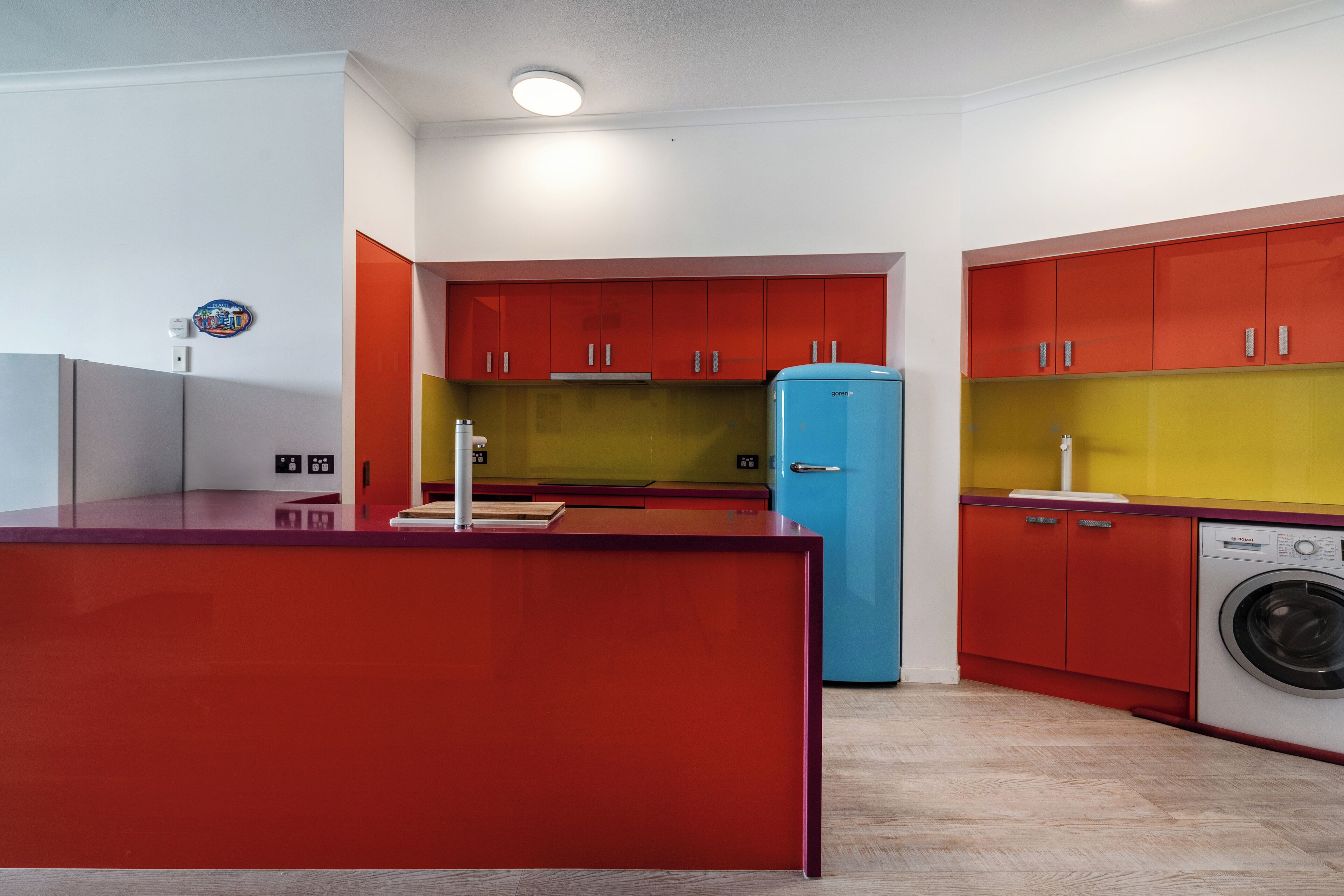 Una cocina compacta con armarios rojos y amarillos, un refrigerador azul y una lavadora.