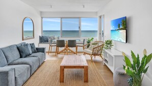 Een moderne woonkamer met uitzicht op de oceaan, een grijze bank, een houten salontafel en een eethoek met stoelen.