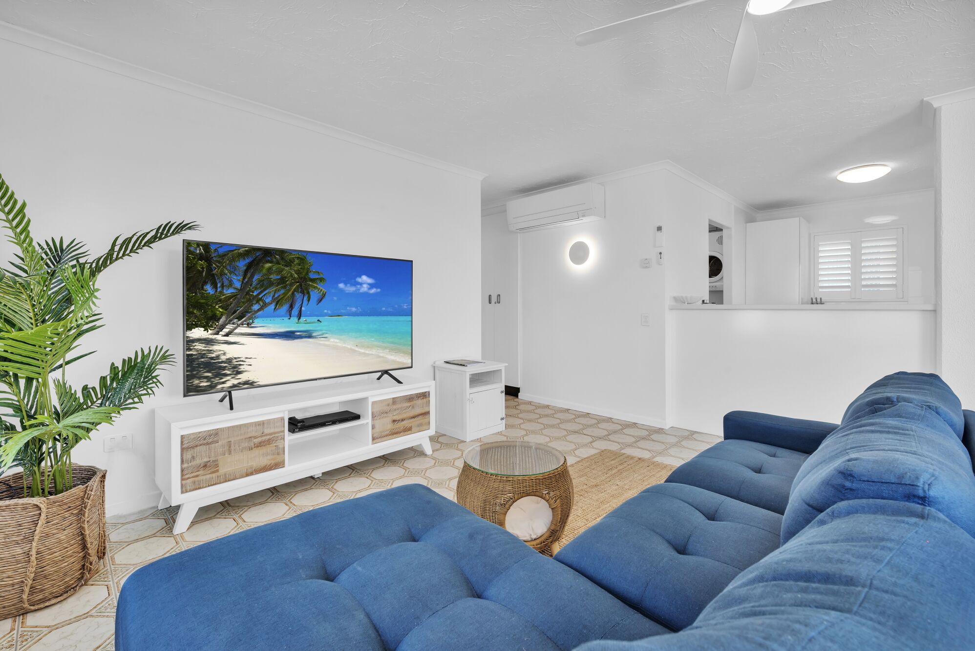 A modern living room with a large flat-screen TV, blue sofas, a white entertainment unit, and a kitchen area with white cabinets and a washing machine.