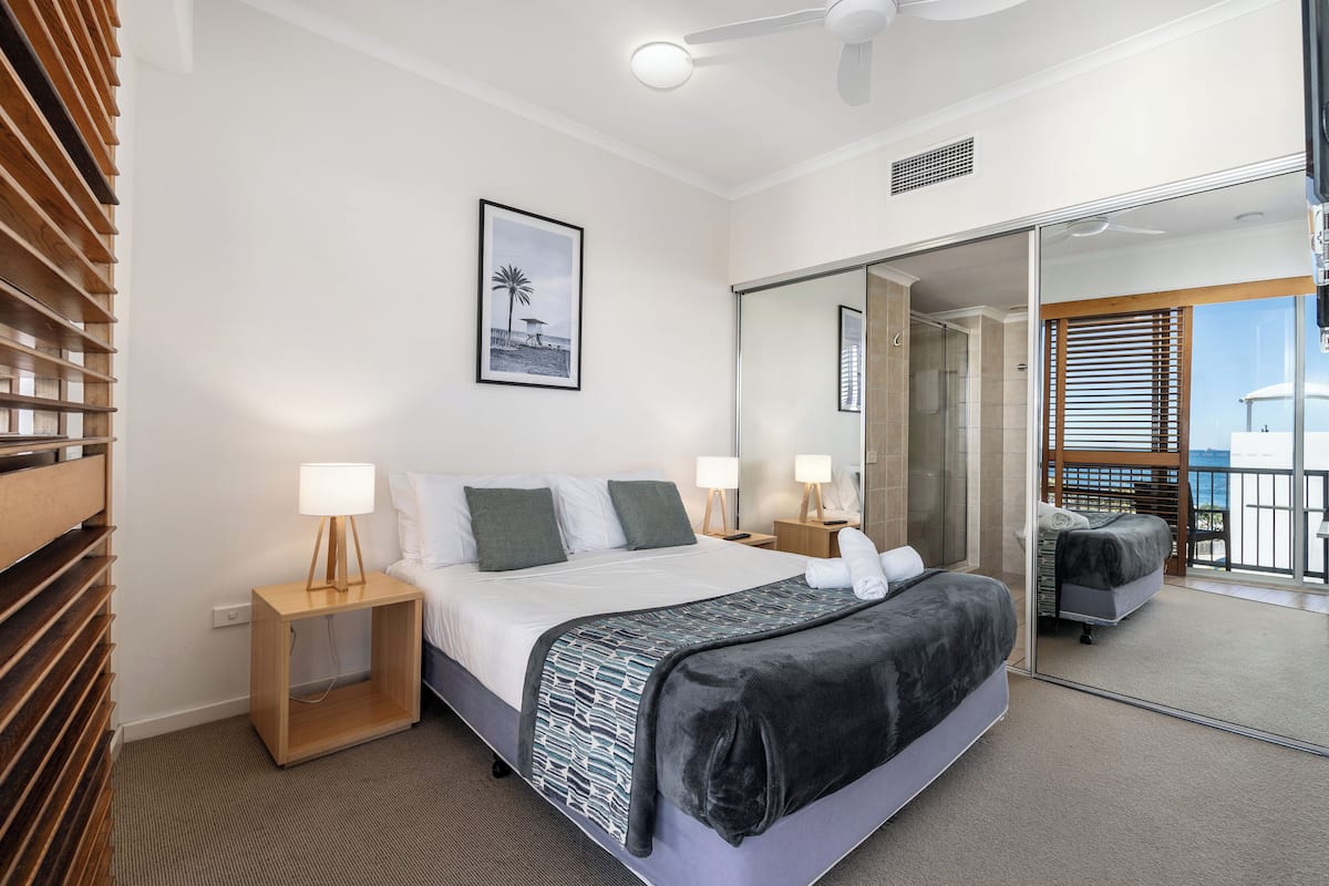 A modern hotel room with a large bed, two bedside tables with lamps, a large mirror, and a view of the balcony.