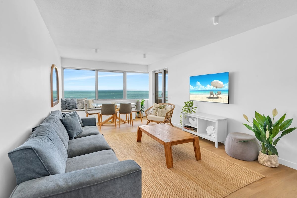 A modern living room with a grey sofa, wooden coffee table, and a view of the ocean through large windows.