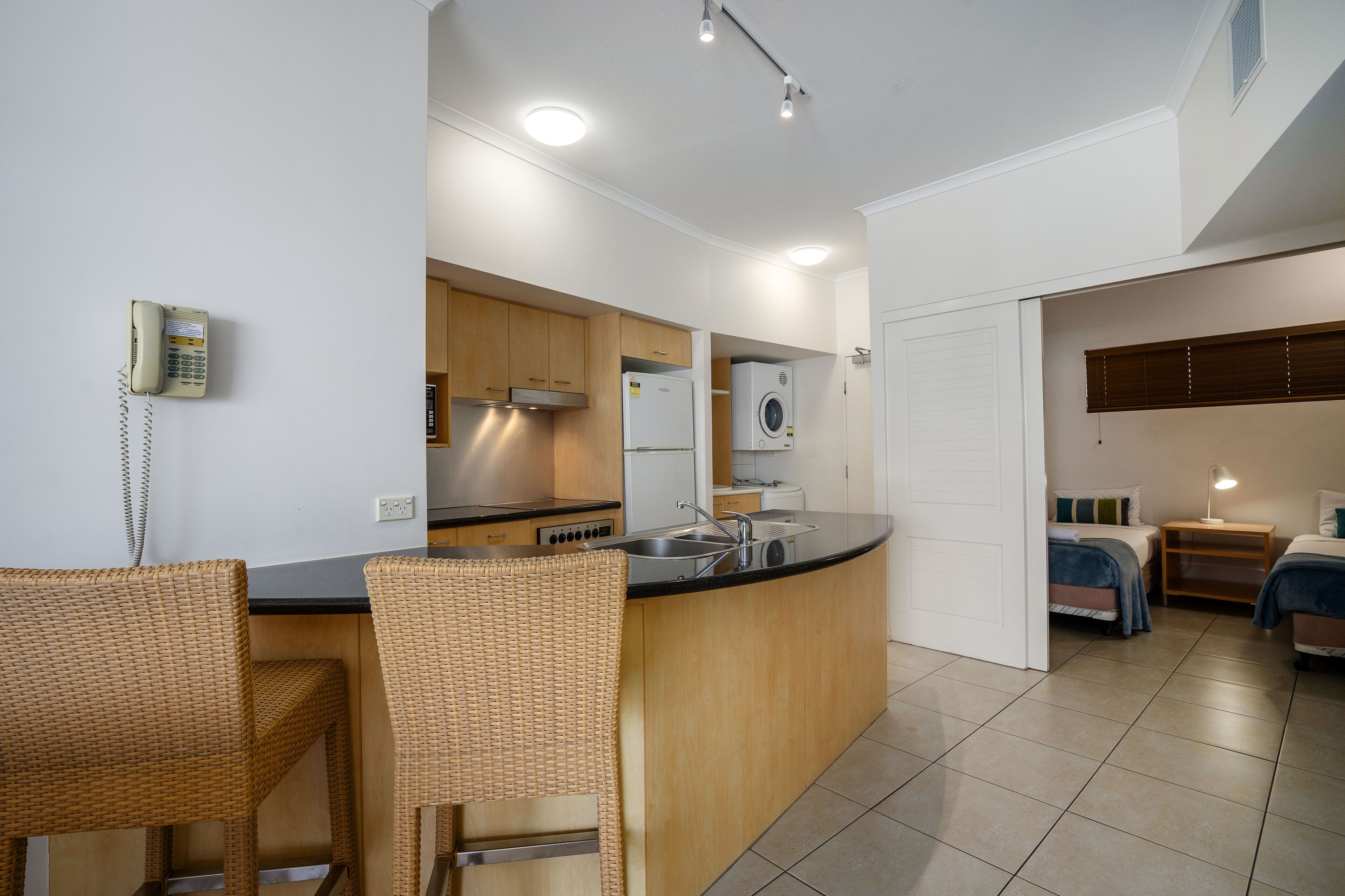 Una cocina compacta con una isla integrada, un comedor con sillas de mimbre y un dormitorio con cama y mesita de noche.