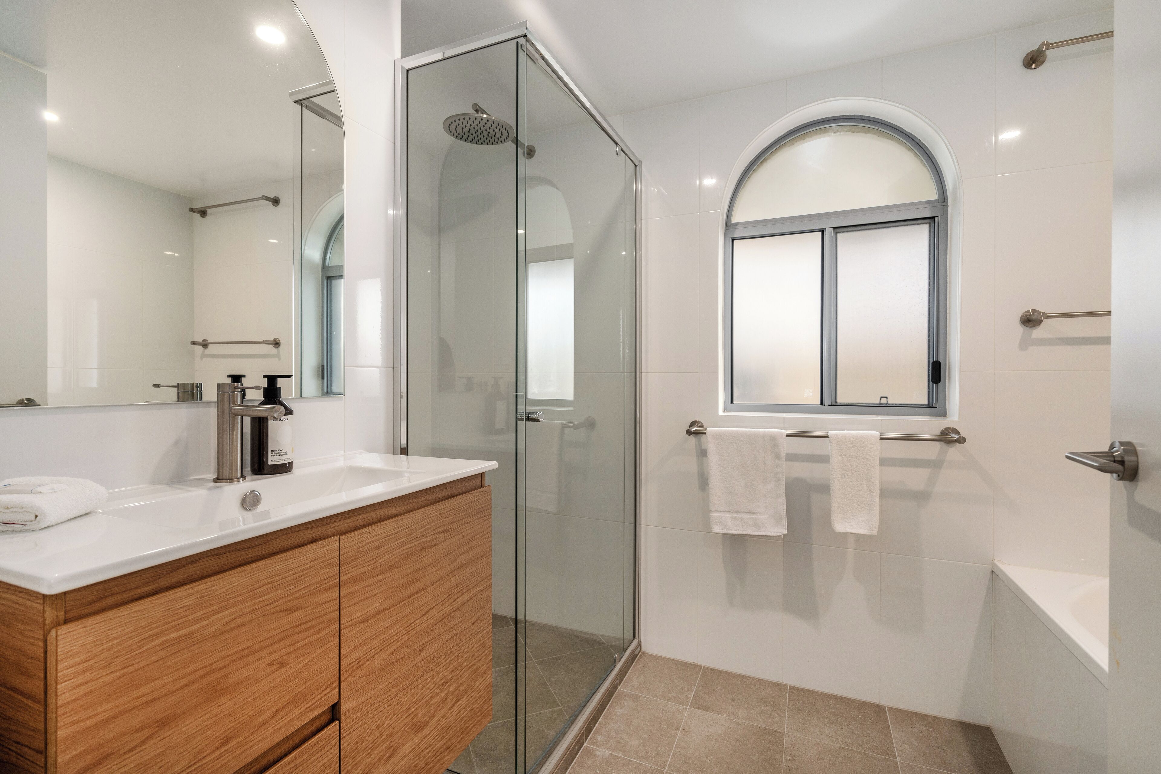 A modern bathroom with a glass-enclosed shower, a wooden vanity, a large mirror, and a window with a frosted glass design.