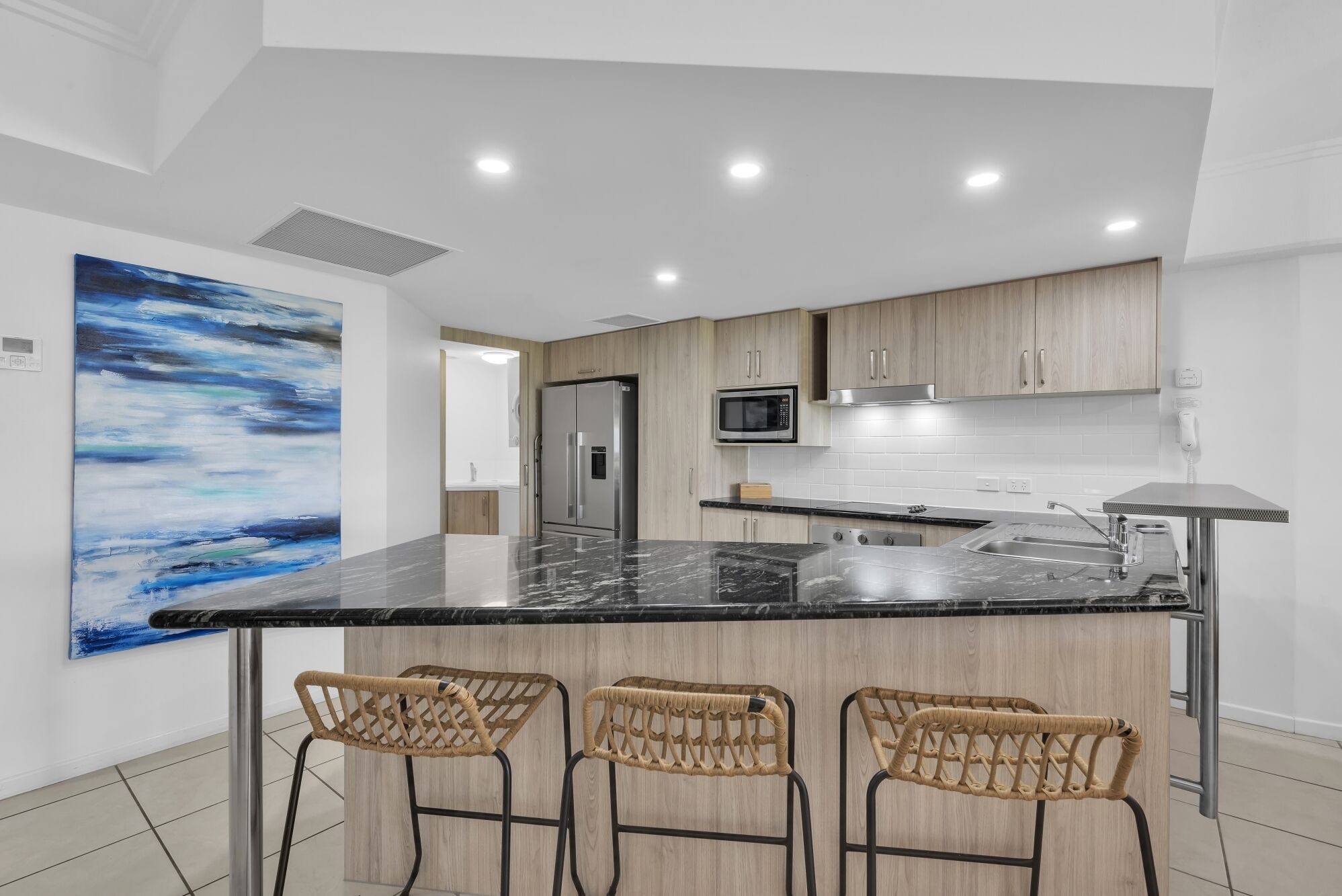 A modern kitchen with a marble countertop, stainless steel appliances, and three wicker bar stools.