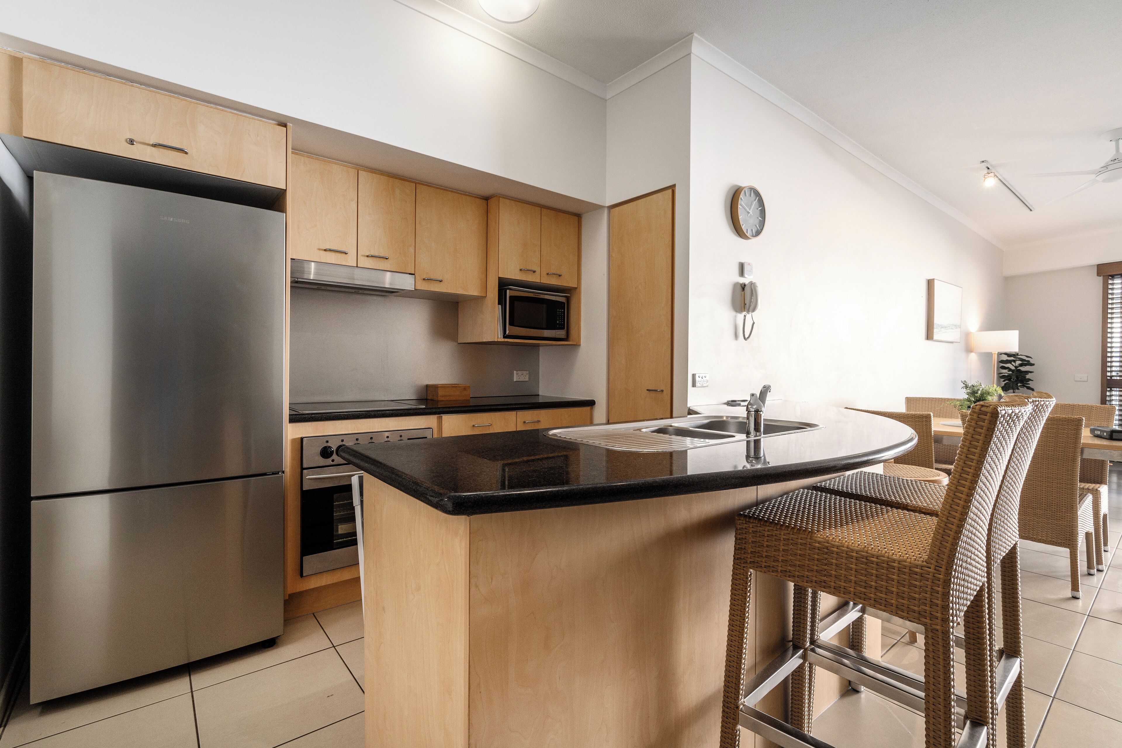 A modern kitchen with a stainless steel refrigerator, microwave, and oven, wooden cabinets, a black countertop island, wicker bar stools, and a dining area with a table and chairs.