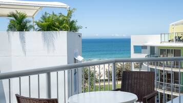 A balcony with a white table and wicker chairs, overlooking a beach and ocean.