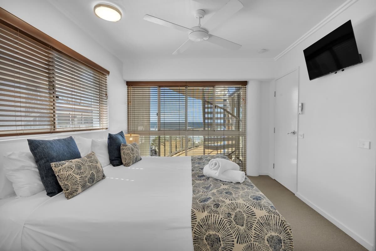 A bedroom with a bed, a ceiling fan, a TV mounted on the wall, and a window with blinds.
