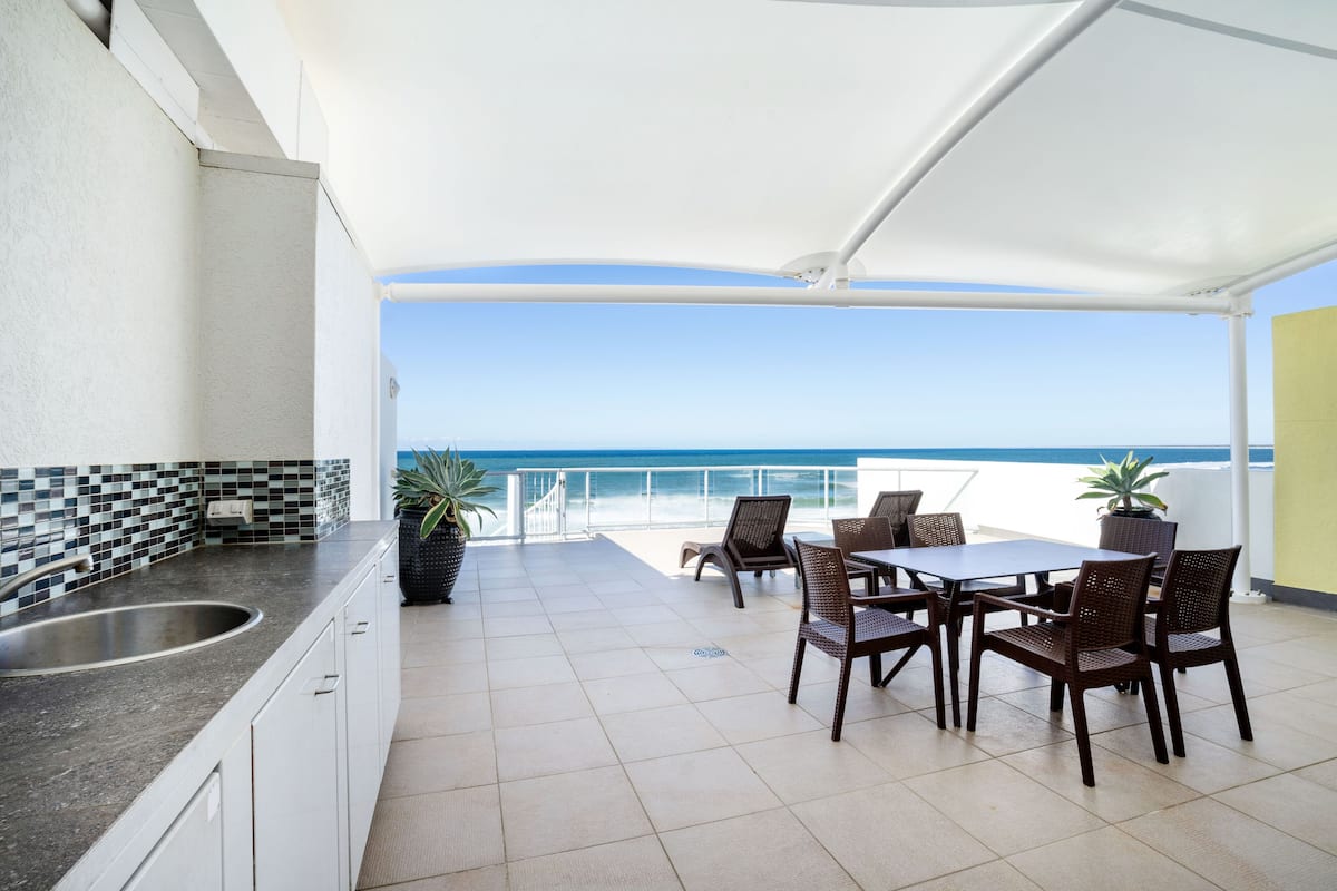 A kitchen with a sink and cabinets, a dining area with chairs and a table, and a view of the ocean.
