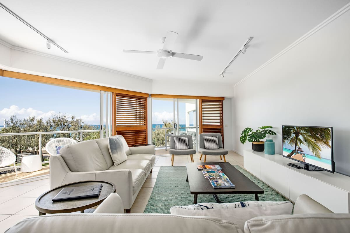 A modern living room with a balcony, white furniture, a flat-screen TV, and a view of the ocean.