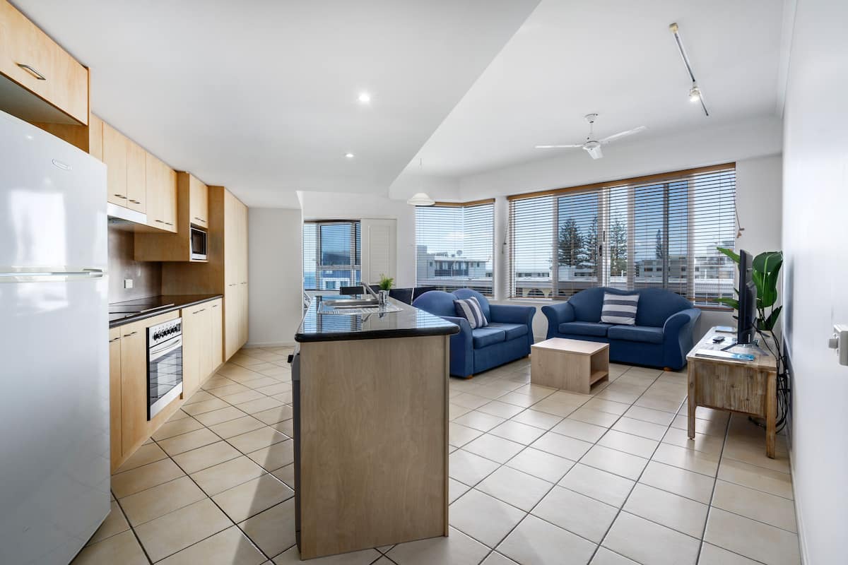 A modern kitchen with a central island, a dining area with a blue sofa, and large windows offering a city view.