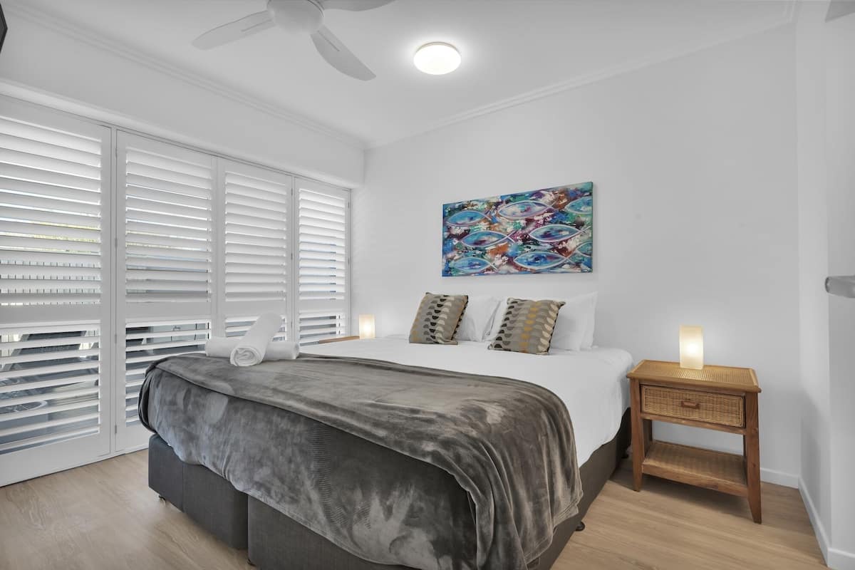 A bedroom with a bed, a ceiling fan, a window with blinds, a wall-mounted artwork, and a bedside table with a lamp.