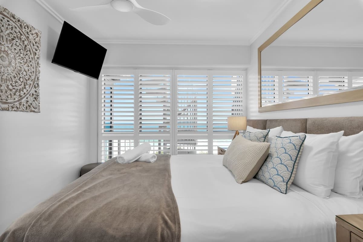 A modern hotel room with a large bed, a flat-screen TV mounted on the wall, and a large window with white blinds.