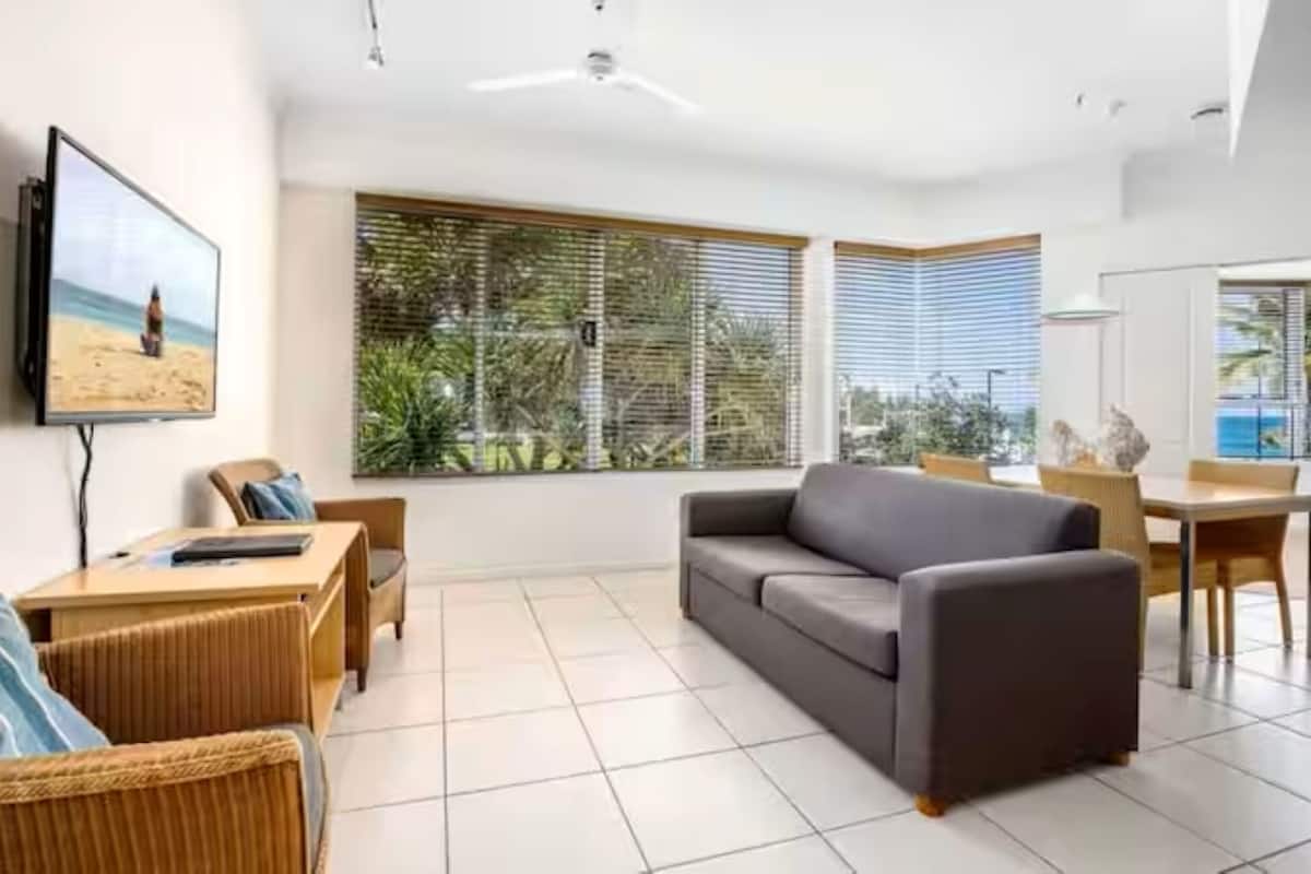 A modern living room with a sofa, a TV, a dining area, and large windows with blinds.