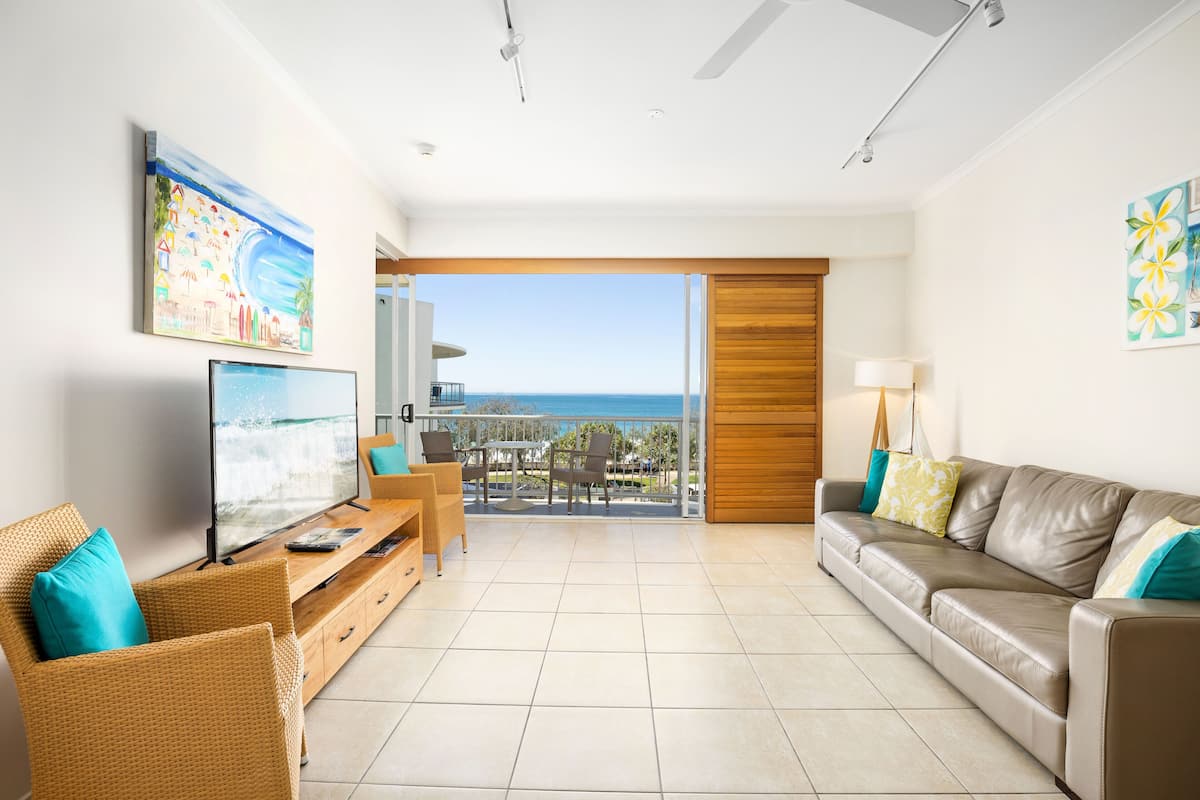 A living room with a television, wicker furniture, a leather sofa, and a view of the ocean through a sliding door.
