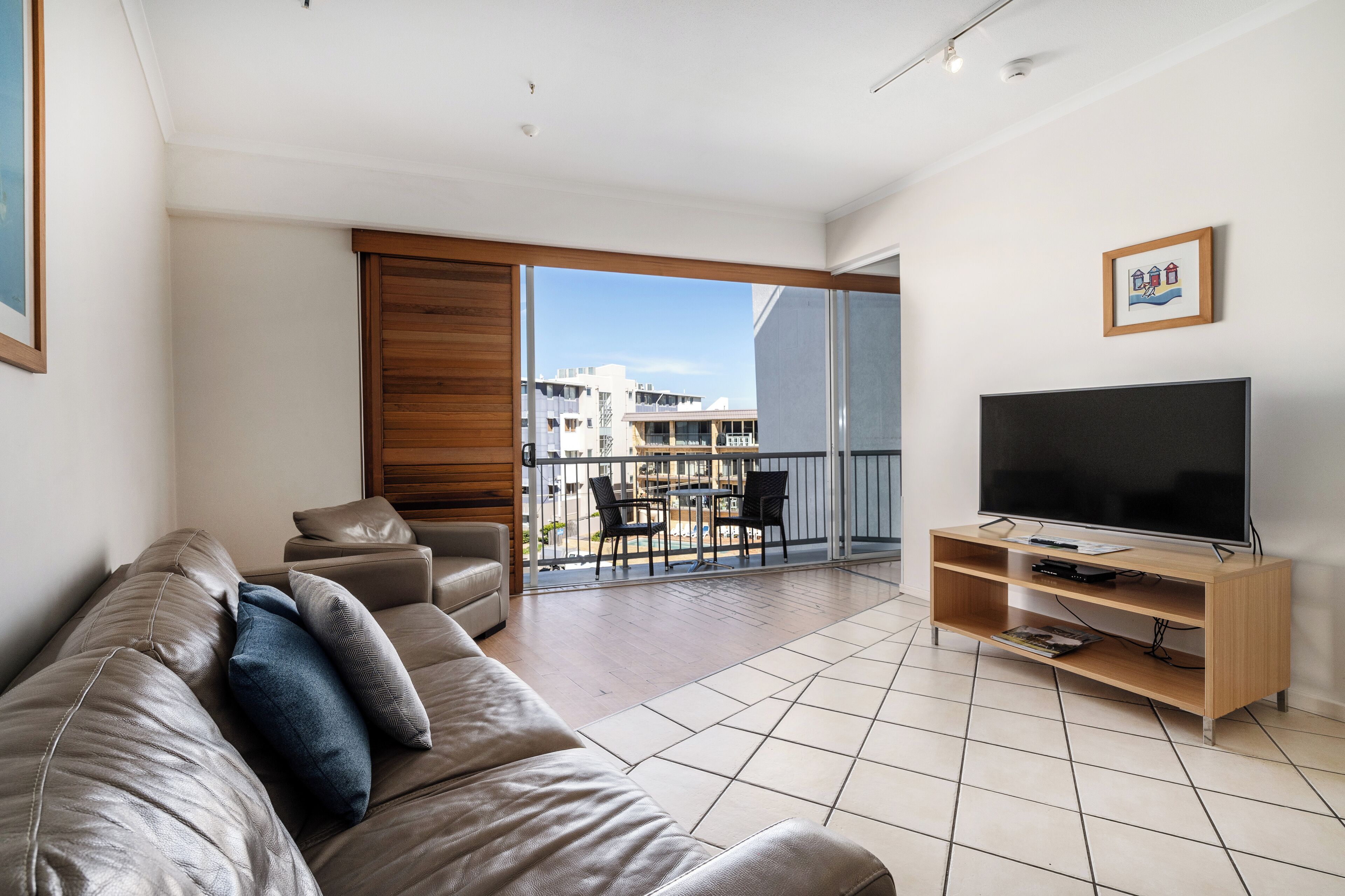 A modern living room with a leather sofa, a flat-screen TV on a wooden stand, and a balcony with a view of buildings.
