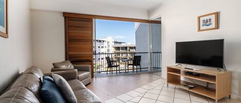 A modern living room with a leather sofa, a flat-screen TV on a wooden stand, and a balcony with a view of buildings.
