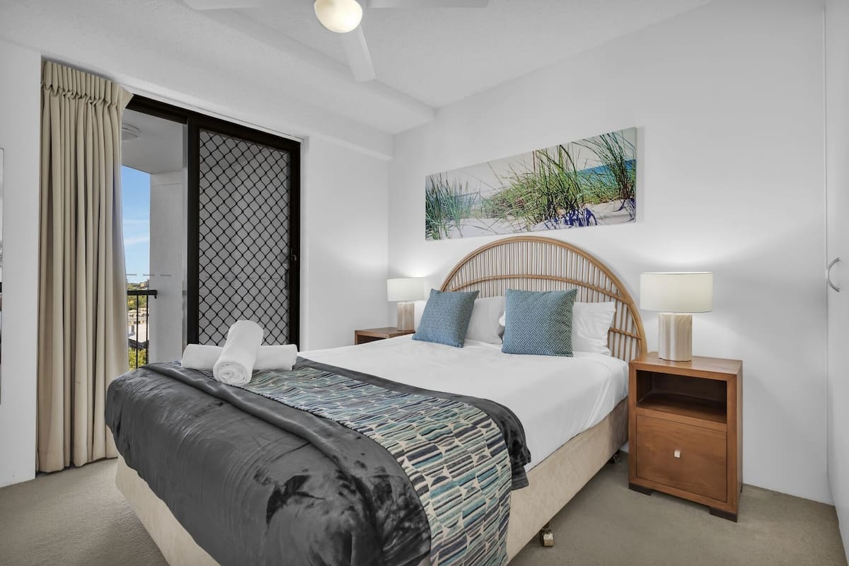 A modern bedroom with a large bed, two bedside tables with lamps, a view through a sliding door, and a beach-themed painting on the wall.