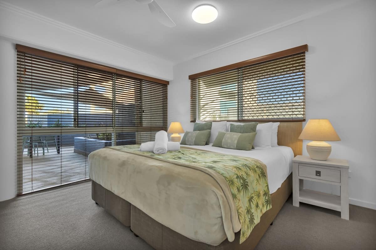 A bedroom with a large bed, two bedside tables with lamps, and a view through floor-to-ceiling windows with blinds.