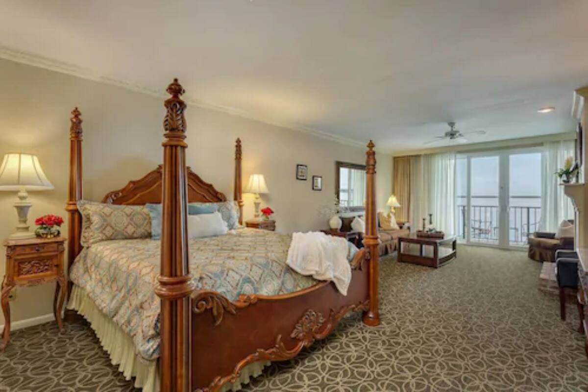 A four-poster bed with ornate wooden carvings, a patterned bedspread, and a matching nightstand with a lamp. The room has a patterned carpet, a small table with a lamp, and a view of the outdoors through large windows.