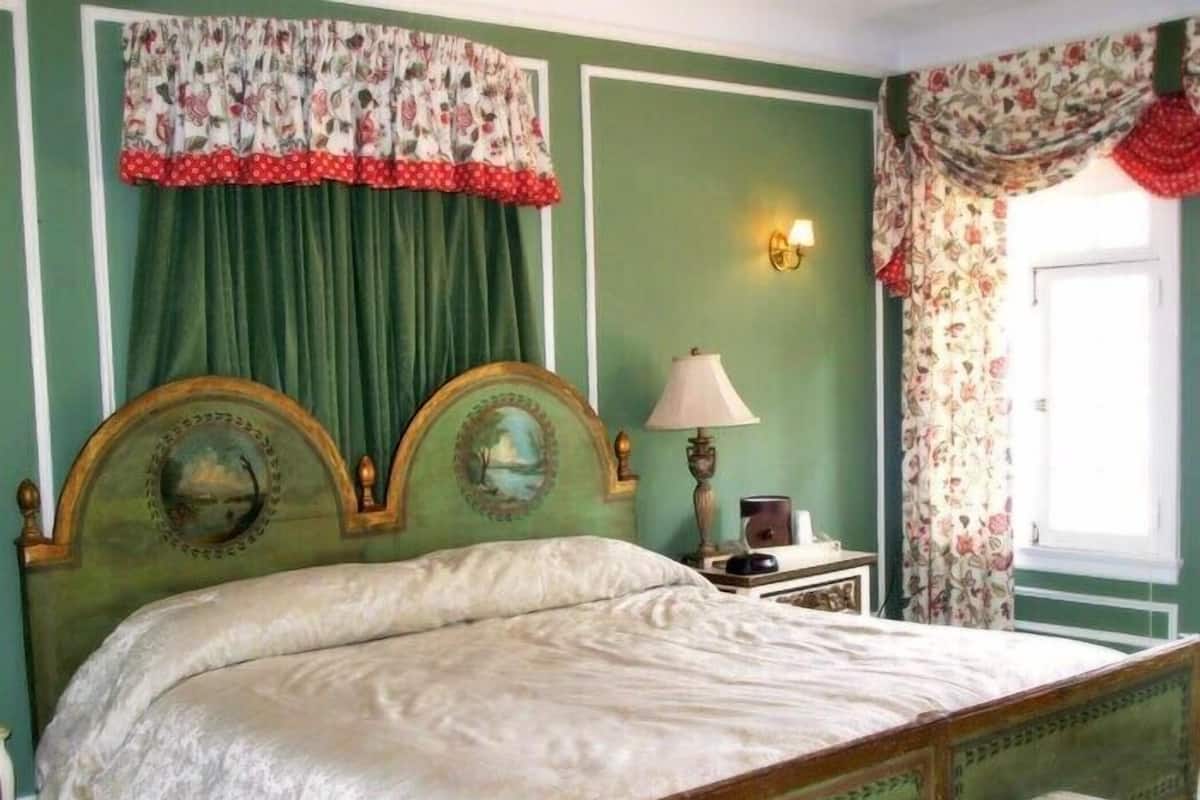 A bedroom with a bed, green walls, floral curtains, and a nightstand with a lamp.