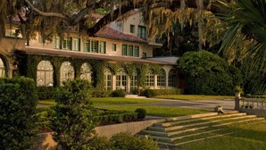A large, historic building with arched windows and a red-tiled roof, surrounded by well-maintained gardens and a paved driveway.