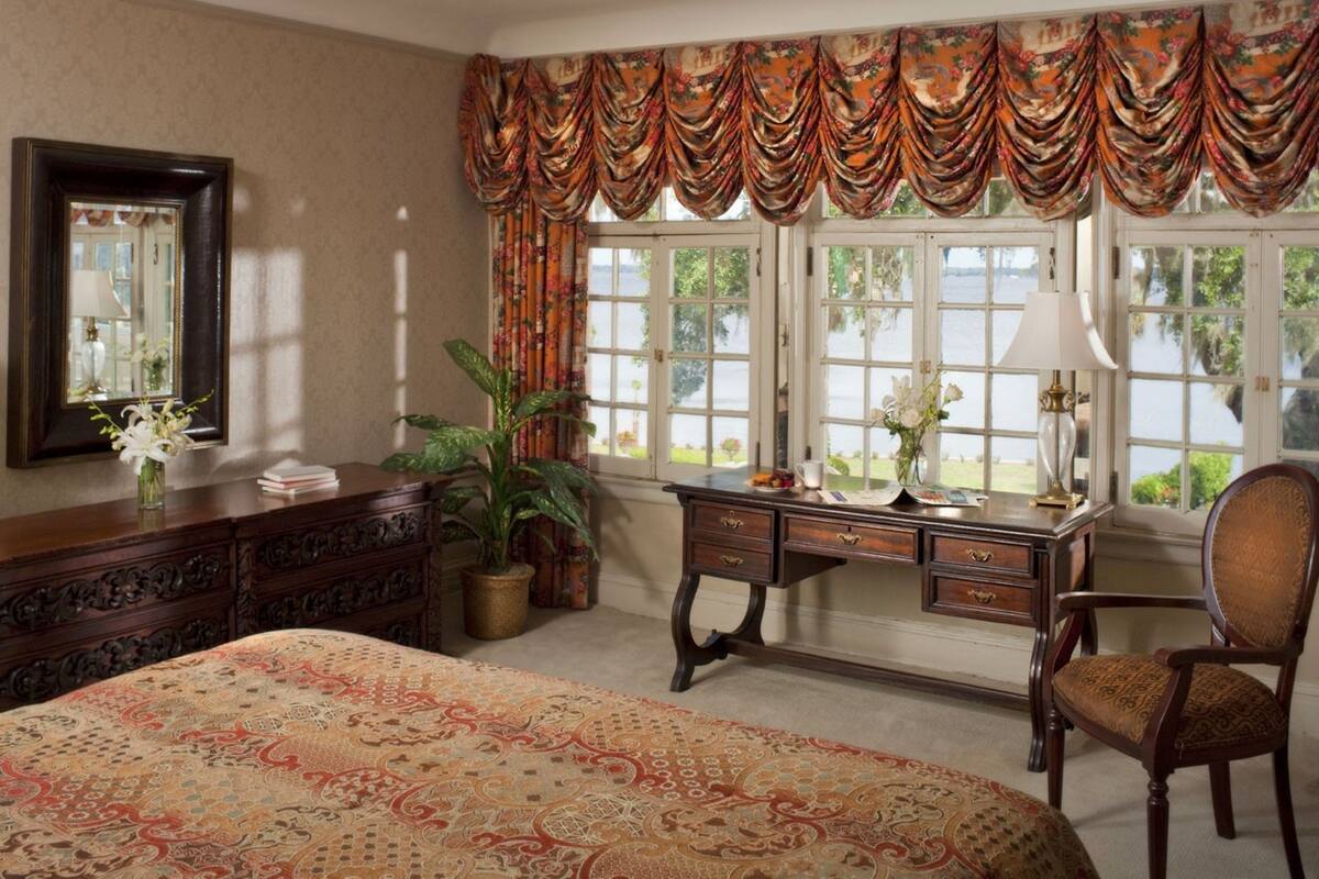 A bedroom with a large bed, a desk with a lamp, a chair, a dresser, and a window with curtains.