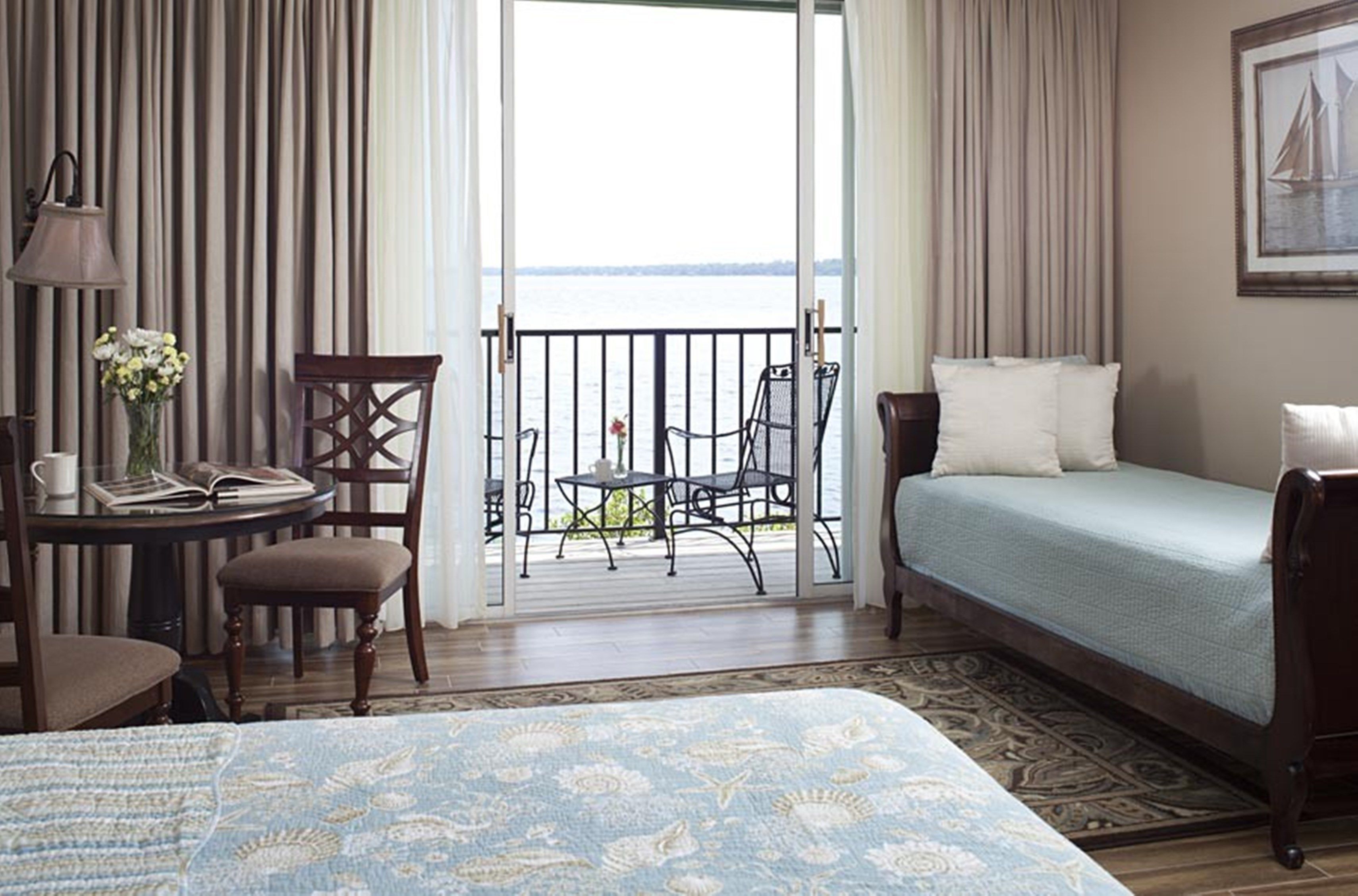 Habitación de hotel con cama, una mesita con una silla, vista al mar desde un balcón y un cuadro en la pared.