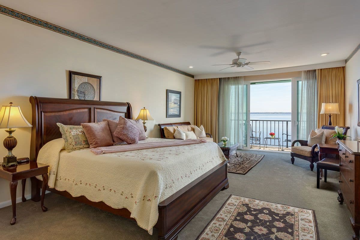 A hotel room with a large bed, two bedside tables with lamps, a view of the sea through a balcony, and a ceiling fan.