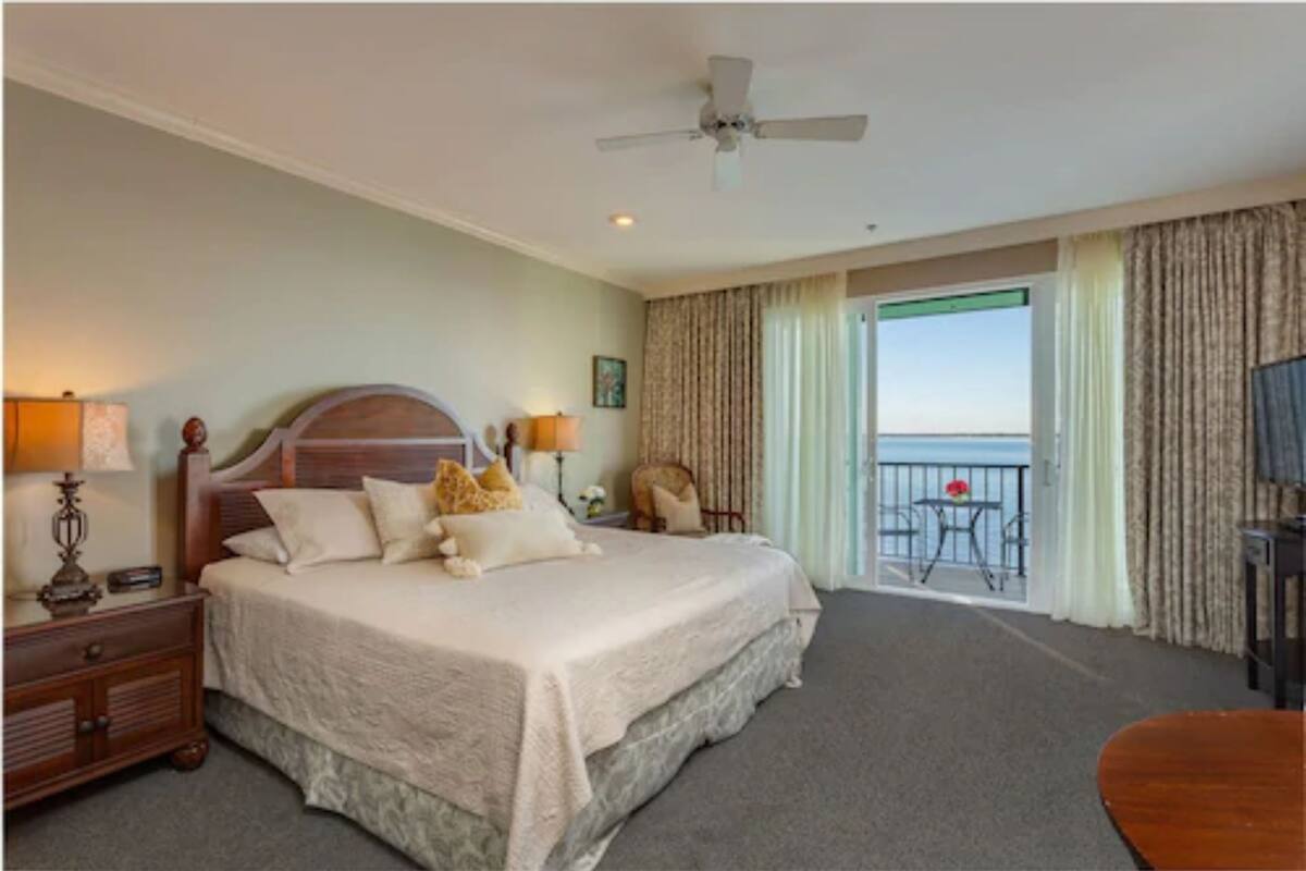 A hotel room with a large bed, a ceiling fan, a balcony with a table and chairs, and a view of the ocean.