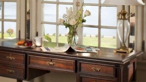 A desk with a lamp, a vase with flowers, and a view of a body of water through a window with curtains.