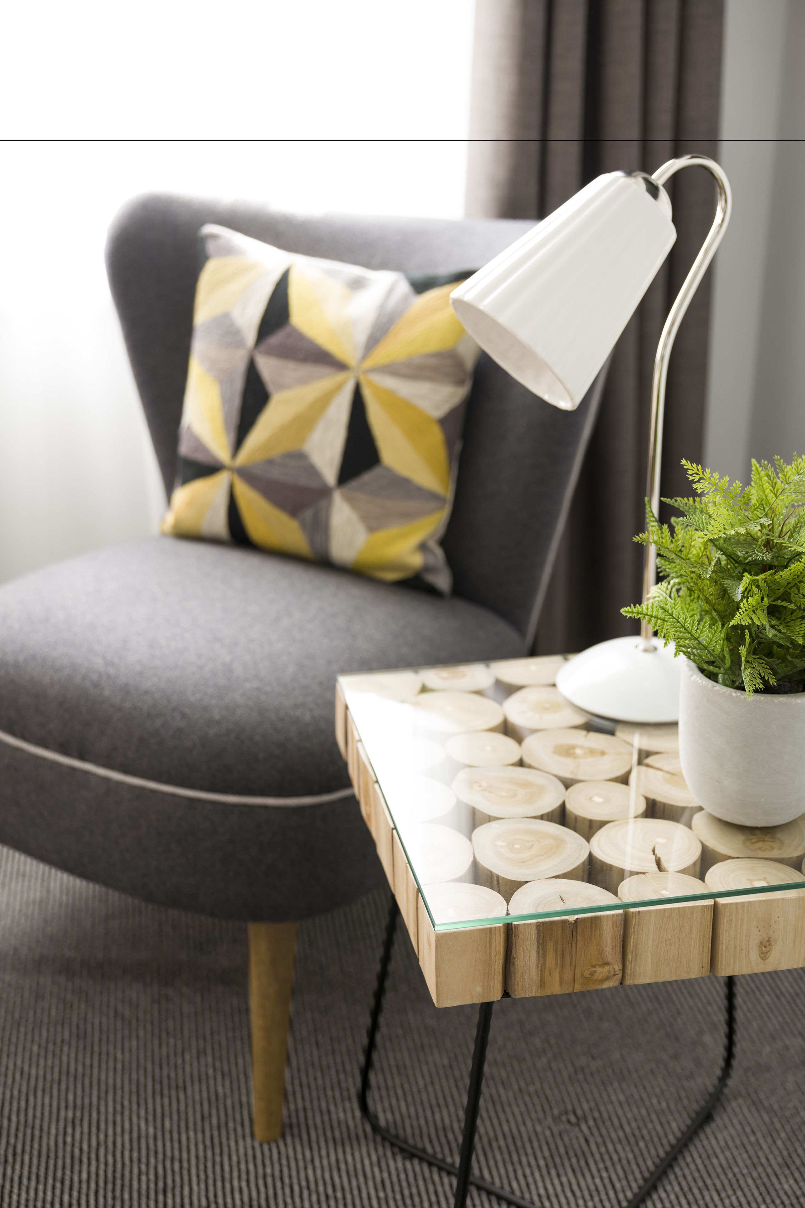 A modern room with a grey sofa, a geometric patterned pillow, a white desk lamp, and a potted plant on a wooden and glass table.