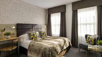 A modern hotel room with a large bed, a desk, and a chair. There is a nightstand with a lamp and a potted plant. The room has large windows with curtains.