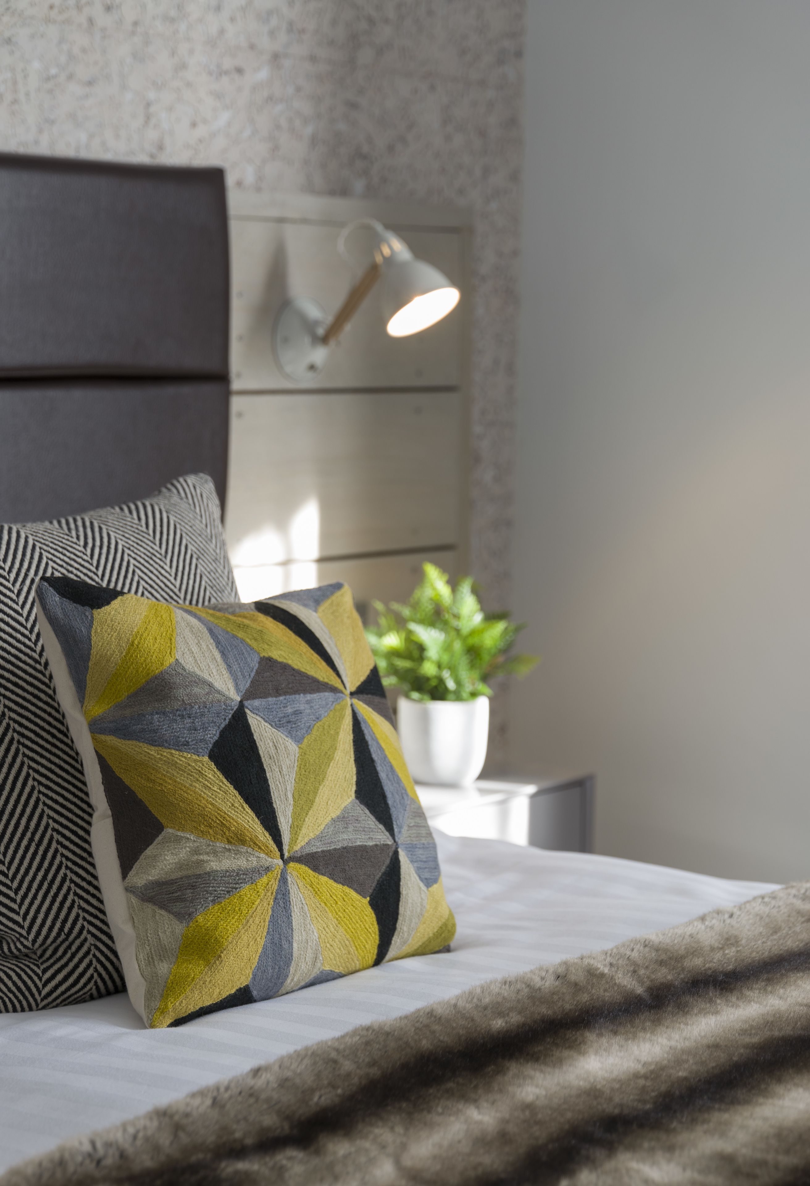 Cama bem arrumada com almofadas estampadas, abajur e planta em vaso.