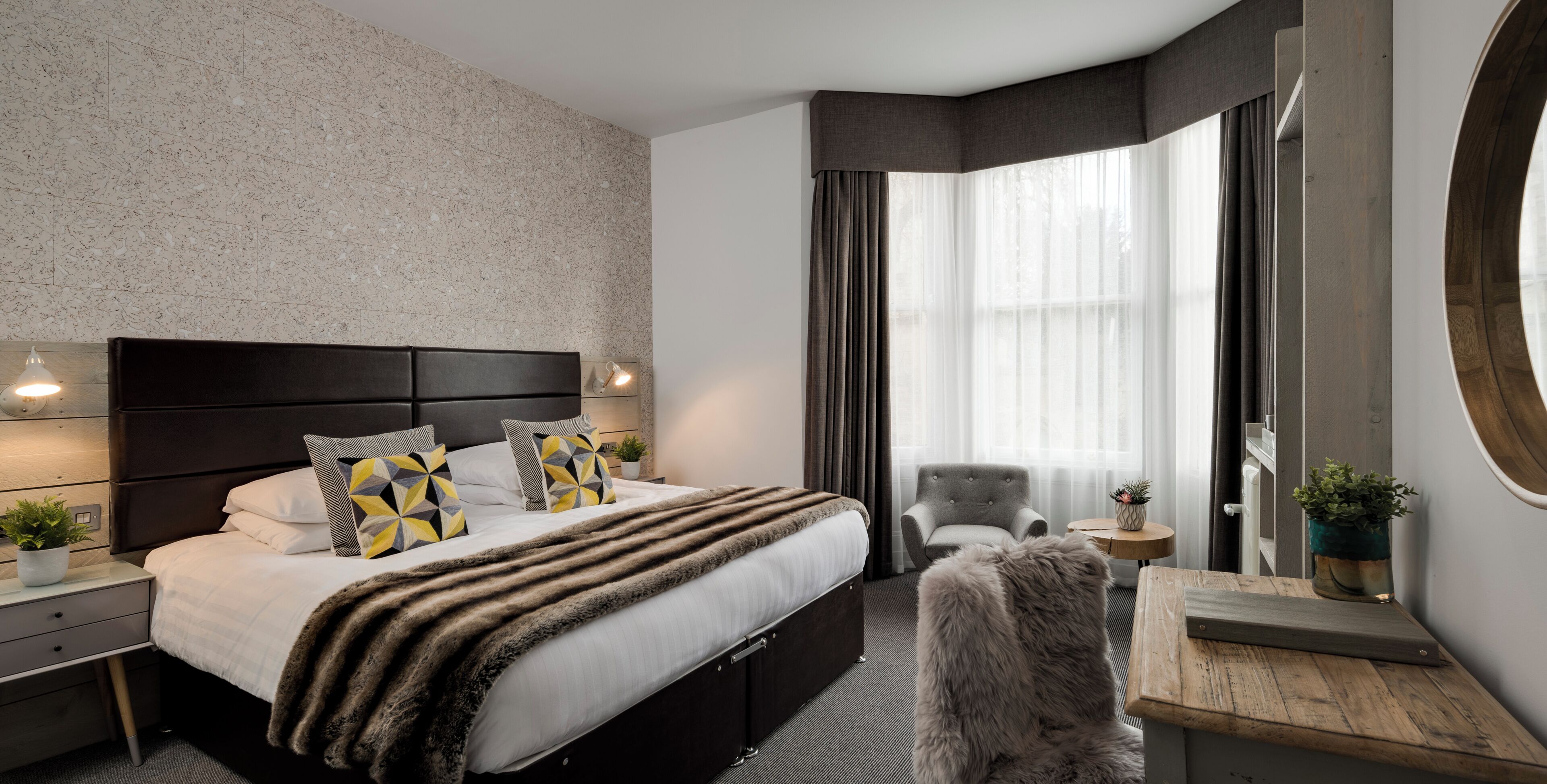 A modern hotel room with a large bed, a grey armchair, a small wooden table, and a round mirror.