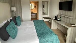 Quarto de hotel com cama, secretária, televisão e casa de banho visível através de uma porta aberta.