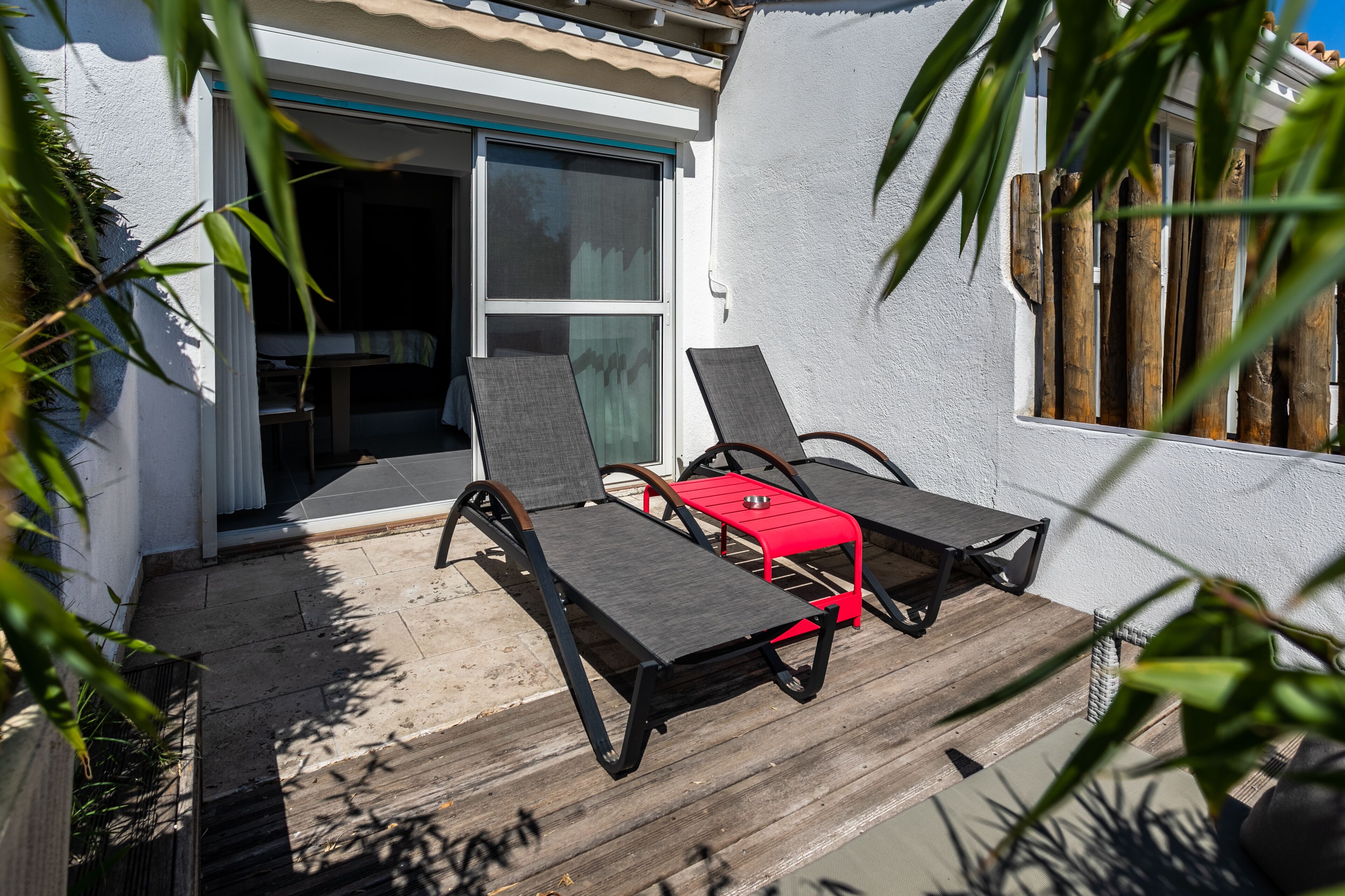 A patio with two lounge chairs and a red table, a sliding glass door, and a white wall.