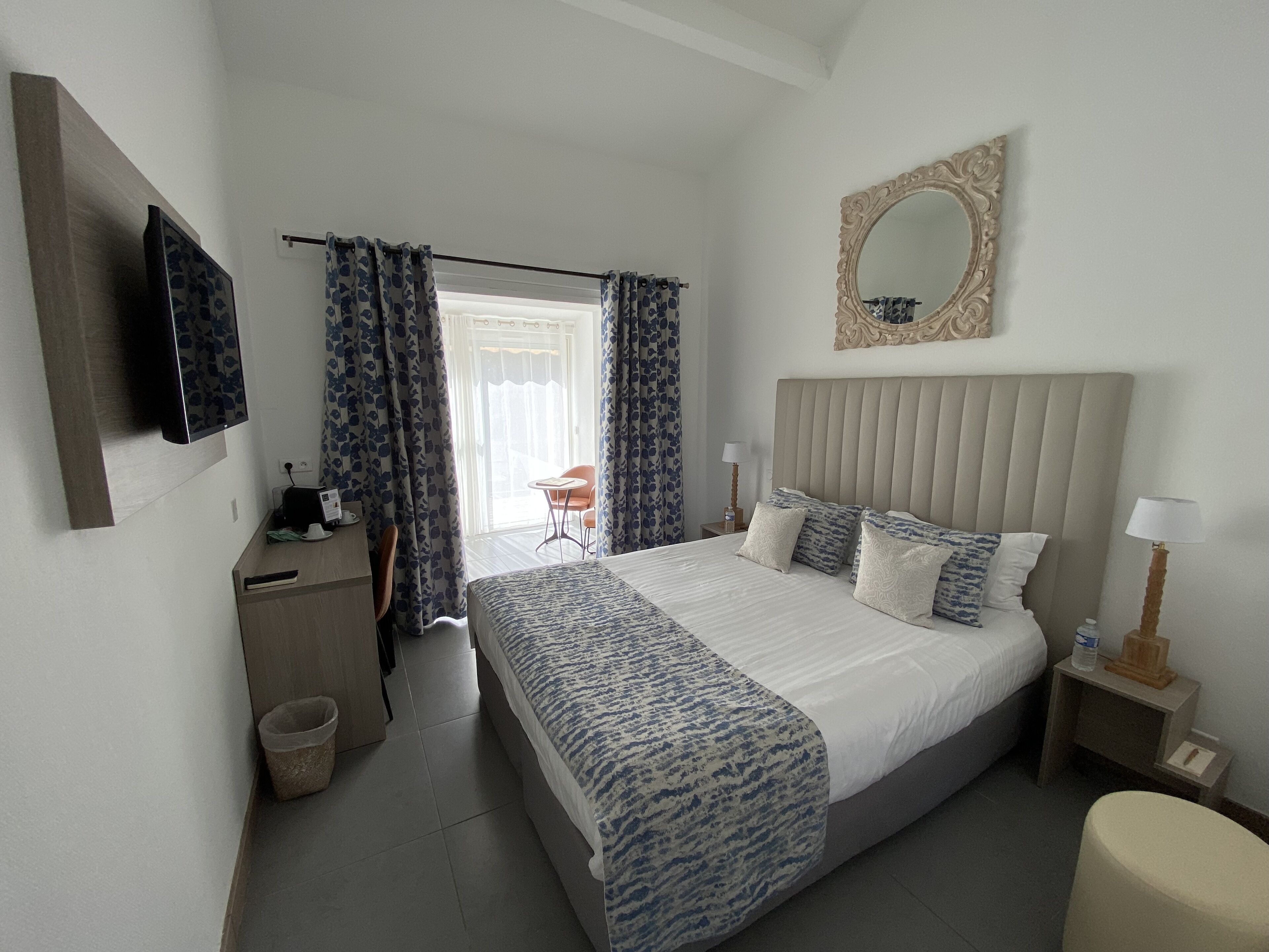 Une chambre d’hôtel avec un lit, un téléviseur fixé au mur, une table de chevet avec une lampe et une vue sur l’extérieur par une fenêtre munie de rideaux.