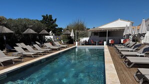 Piscina com espreguiçadeiras e guarda-sóis, cercada por árvores e um edifício com área de estar ao ar livre.