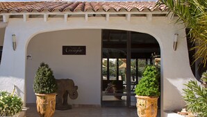 Entrada de um edifício com telhado telhado, placa indicando 'Casa de Oceango' e duas grandes plantas em vasos.