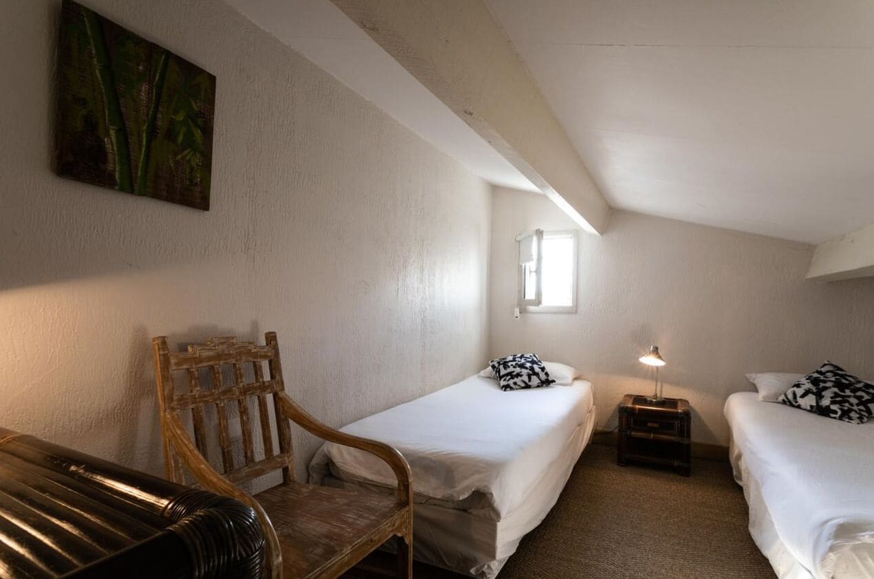 Une petite chambre accueillante, avec deux lits simples, une chaise en bois, une petite table avec une lampe et un tableau au mur.