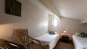 Une petite chambre accueillante, avec deux lits simples, une chaise en bois, une petite table avec une lampe et un tableau au mur.