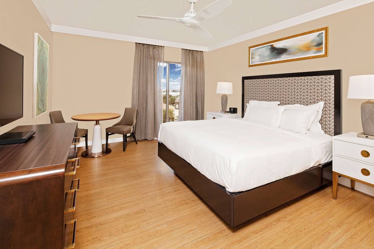 Ein Hotelzimmer mit einem großen Bett, einem Schreibtisch mit Stuhl, einem kleinen runden Tisch und einem Deckenventilator.