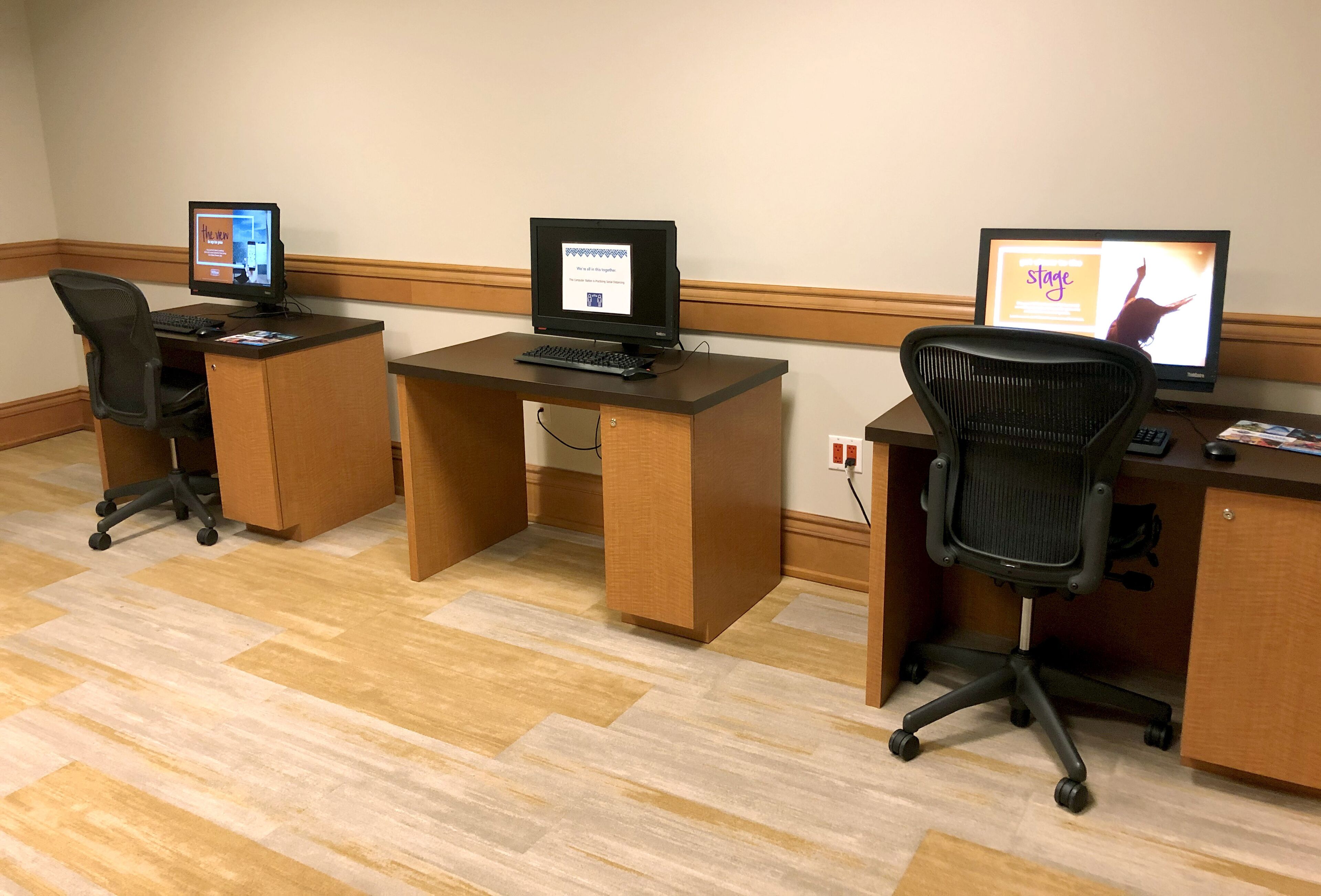 A hotel room with three desks, each with a computer and a chair.