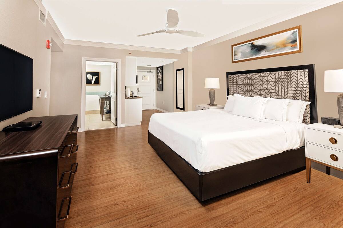 Ein modernes Hotelzimmer mit einem großen Bett, einem Schreibtisch mit Schubladen, einem Flachbildfernseher, einem Deckenventilator und einem Gemälde an der Wand.