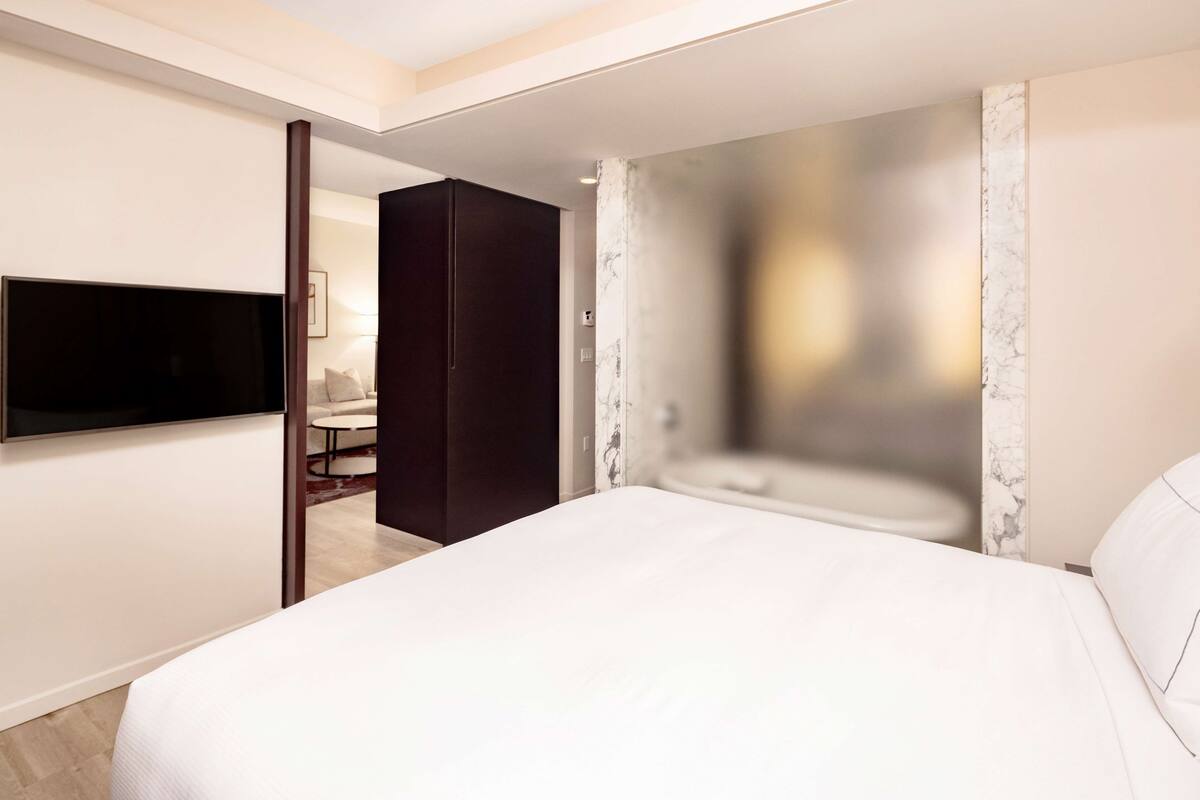A modern hotel room with a large bed, a flat-screen TV mounted on the wall, and a dark-colored wardrobe.