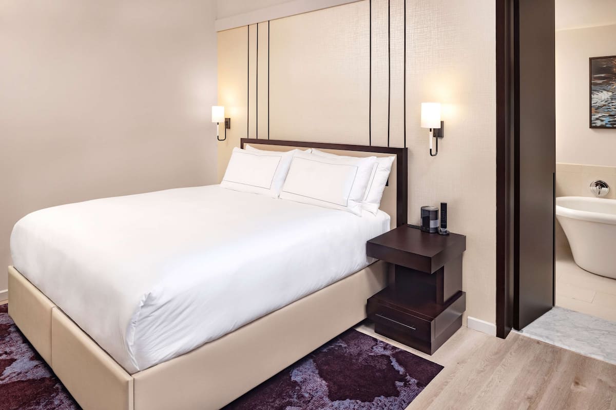 A neatly made bed with white linens, a dark nightstand with a lamp, and a bathroom with a bathtub and a toilet visible.