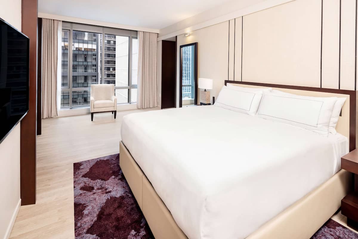 A hotel room with a large bed, a TV, a chair, and a view of the city through large windows.